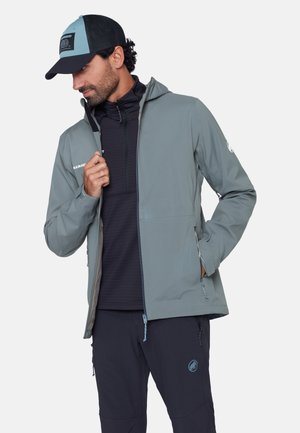 Mann trägt eine hellgraue Kapuzenjacke über einem schwarzen Reißverschluss-Shirt und dunkler Hose, zieht mit einer Hand den Reißverschluss der Jacke hoch.