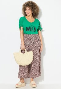 Grüne T-Shirt mit dem Aufdruck "Stay WILD" in Schwarz, kombiniert mit braunen weit geschnittenen Hosen im Leopardenmuster; hält eine runde gewebte Strohtasche; trägt sandfarbene Plateausandalen.