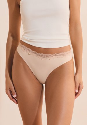 Mujer con ropa interior beige con encaje y una camiseta de tirantes blanca con canalé, vista desde la mitad del torso hasta la parte superior de los muslos.