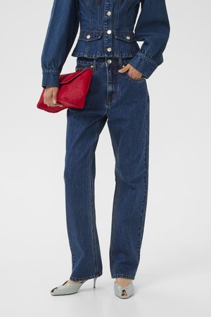 Woman wearing dark blue denim jacket and jeans, holding a red clutch, with light gray high-heeled open-toe shoes.