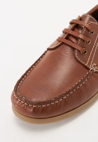 Chaussure style mocassin en cuir marron avec coutures blanches visibles, laçage à l’avant et semelle en caoutchouc marron clair sur fond blanc.