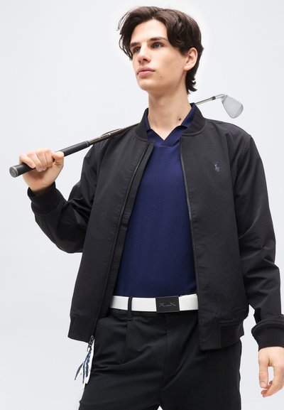 Νεαρός άνδρας με μπλε ζακέτα και μπλε πόλο κρατάει μπαστούνι γκολφ στον ώμο, φοράει μαύρο παντελόνι και λευκή ζώνη πάνω σε απλό φόντο.