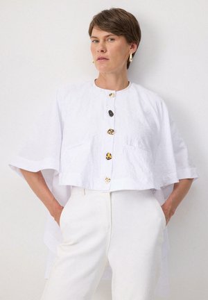 Haut blanc, crop top à boutons avec des manches larges, présentant des boutons décoratifs en or et noir. Tissu lisse avec une texture douce.