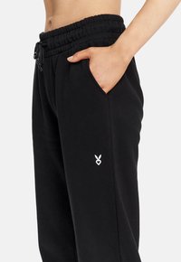 Schwarze Jogginghose mit einem Zugbandbund, Seitentaschen und einem weißen gestickten Logo am Oberschenkel. Weicher Stoff mit lockerer Passform.