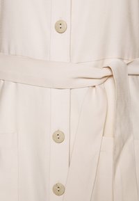 Šaty krémové barvy se zapínáním na knoflíky vpředu, látkovým páskem, dvěma předními kapsami a hladkou texturou. Knoflíky jsou kulaté s malými otvory.