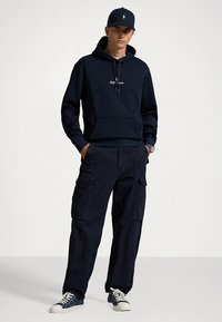 Sweatshirt azul-marinho com bolso frontal e capuz ajustável, combinada com calças cargo azuis-marinho com bolsos laterais e ténis azuis-marinho com solas brancas.