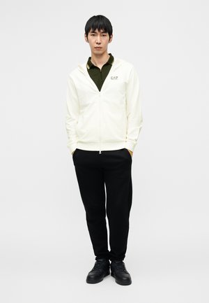 Sudadera blanca con cremallera y capucha, sobre una camisa polo verde oscuro. Pantalones negros ajustados y zapatillas negras completan el conjunto.