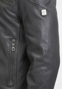 Giacca in pelle nera con finitura testurizzata, dotata di una tasca laterale con zip e un patch con logo sulla spalla. Design resistente e aderente.
