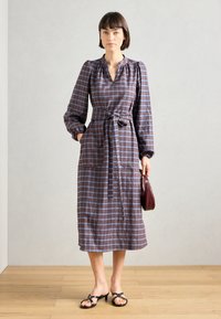 Rutet knelengt kjole i brun, blå og hvit, med belte i midjen, puffermer og sidelommer. Kombinert med svarte sandaler med remmer.