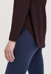 Bordeauxrotes Langarmoberteil mit abgerundetem Saum, kombiniert mit blauen Leggings; glatte Textur, lockere Passform und sichtbare Steppnähte.