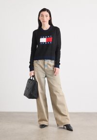 Une femme se tient devant un fond neutre, portant un pull noir avec le logo "TOMMY JEANS", associé à un jean léger et ample, et tenant un sac à main noir.