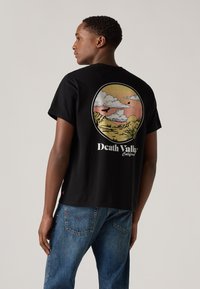 Tricou negru din bumbac, cu un grafic circular pe spate, reprezentând nori și munți în culori pastelate, și textul "Death Valley" sub design.