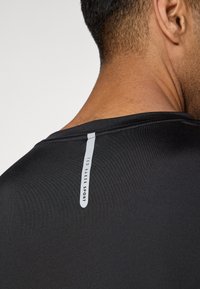 Black athletic shirt, smooth texture, rounded neckline, reflective tag on the back with "TED BAKER SPORT" branding, lightweight material.