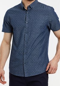 Homme portant une chemise bleu marine à manches courtes, boutonnée, avec de petits motifs géométriques blancs et une poche poitrine.