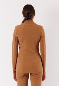 Blazer marrone aderente con maniche lunghe, texture liscia, spalle strutturate e orlo curvo, abbinato a pantaloni aderenti coordinati.