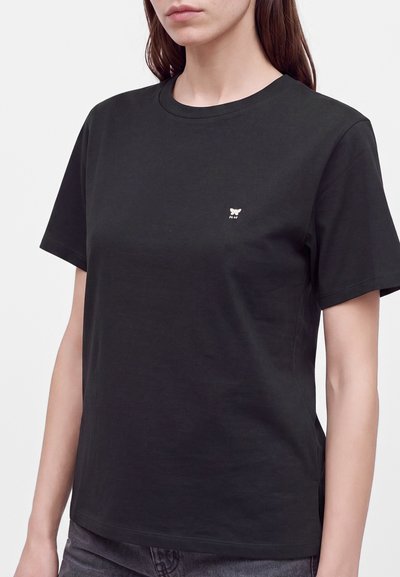 Femme portant un t-shirt noir uni à manches courtes avec un petit logo papillon blanc sur la poitrine gauche.
