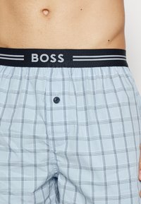 Boxers en tartan bleu clair avec une ceinture élastique noire arborant le logo "BOSS". Comprend une fermeture à bouton et un détail de motif en damier.