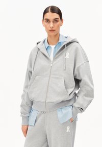 Grauer Kapuzenpullover mit Reißverschluss aus weichem Stoff, mit einer Fronttasche und weißem Logodetail. Dazu ein hellblaues Hemd darunter und passende Hose.