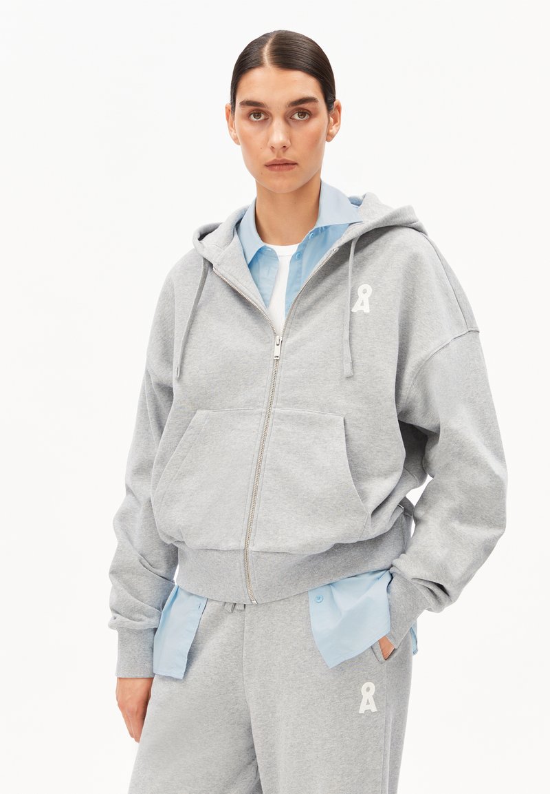 Grauer Kapuzenpullover mit Reißverschluss aus weichem Stoff, mit einer Fronttasche und weißem Logodetail. Dazu ein hellblaues Hemd darunter und passende Hose.