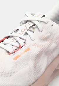 Zapatilla deportiva blanca con tejido texturizado, que presenta sutiles acentos en naranja y rosa, cordones redondos y una suela acolchada con un patrón geométrico.