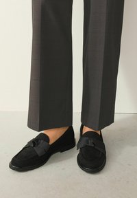 Schwarze Wildleder-Loafers mit flacher Sohle und einer dekorativen Schleife vorne, kombiniert mit grauen karierten Hosen.