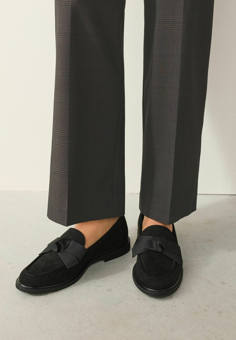 Black suede loafers with a flat sole and a decorative bow on the front, paired with grey plaid trousers.