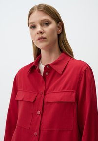 Chemise rouge en tissu tissé, avec col, ouverture à boutons à l'avant et deux poches poitrine. Finition texturée et détails de couture visibles.