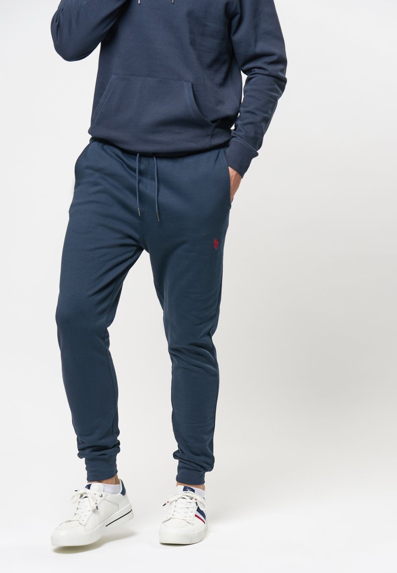 Marineblauwe hoodie met een voorzak, gecombineerd met bijpassende joggers, met een geborduurd logo en een tailleband met trekkoord. Witte sneakers.