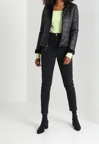 Svart puffjacka med dragkedja, utrustad med en svart fällkrage över en neongrön långärmad topp, kombinerad med högmidjade svarta jeans och ankelstövlar.