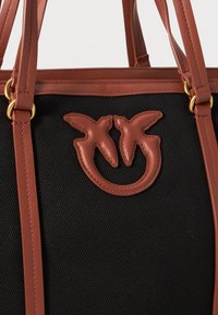 Zwarte canvas totebag met bruin leren accenten. Beschikt over een onderscheidend logo met dubbele vogel en gouden hardware. Dubbele handvatten voor het dragen.