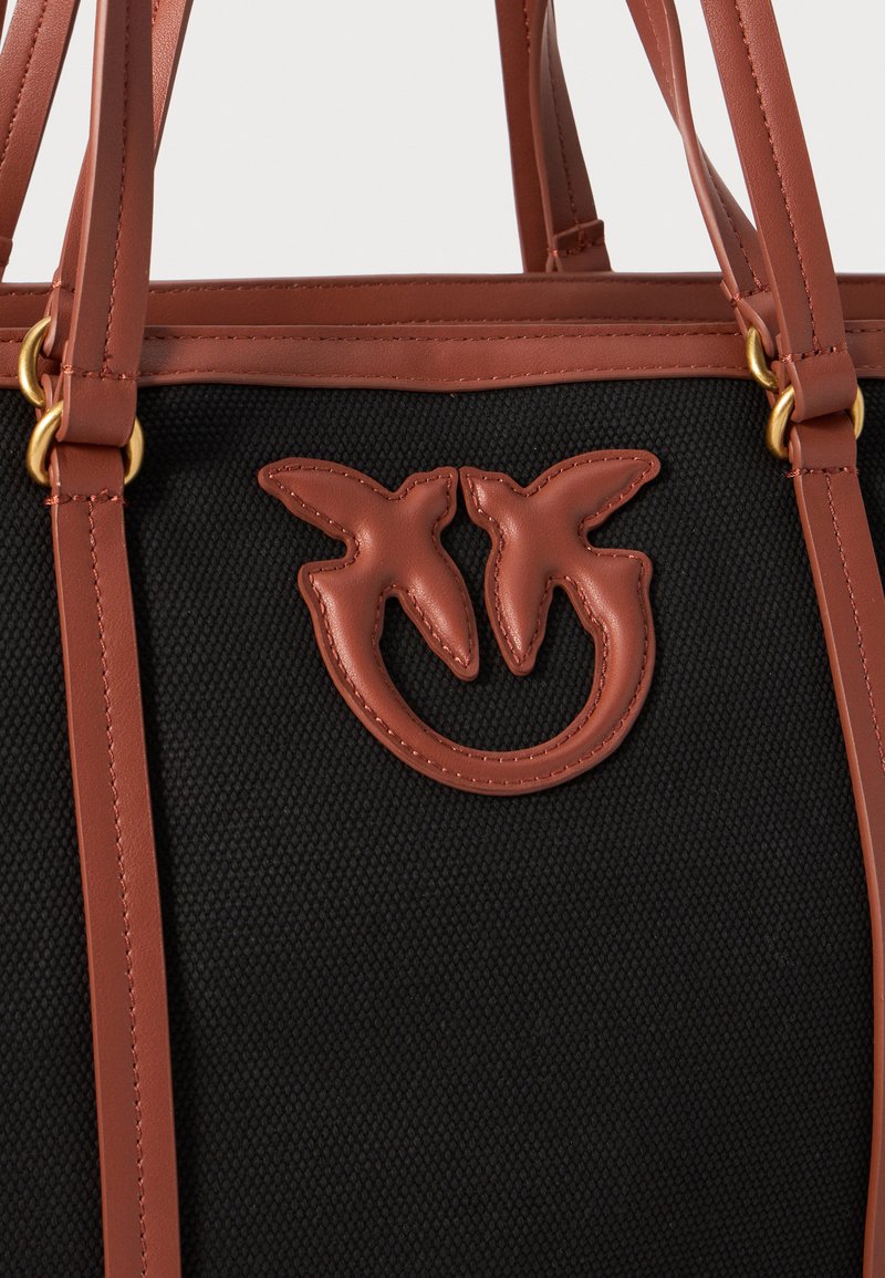 Bolsa de tela negra con detalles de cuero marrón. Presenta un distintivo logo de dos aves y accesorios en tono dorado. Doble asa para llevar.