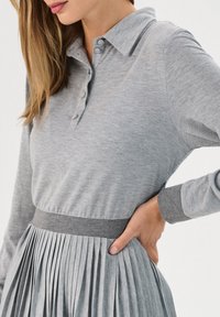 Szary longsleeve polo z zapięciem na guziki, zestawiony z plisowaną spódnicą. Obie sztuki odzieży mają miękką teksturę i dopasowane odcienie szarości.
