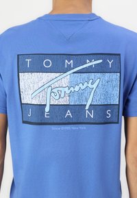 Mėlyna medvilninė marškinė su grafiniu spausdinimu, kuriame užrašas "TOMMY JEANS" baltos ir šviesiai mėlynos spalvos, apjuostas tamsiai mėlynos ir šviesiai mėlynos juostomis.