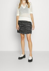 Vaaleanharmaa, lyhythihainen, V-pääntie neulepaita, jossa on avonainen verkkomuotoilu, yhdistettynä mustaan tweed-hameeseen, jossa on kirjaillut reunat ja kultaiset napit.