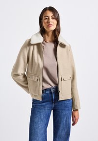 Žena v béžové zipové bundě s bílým fleecovým límcem, s mauve tričkem a modrými džínami, stojí proti jednoduchému bílému pozadí.