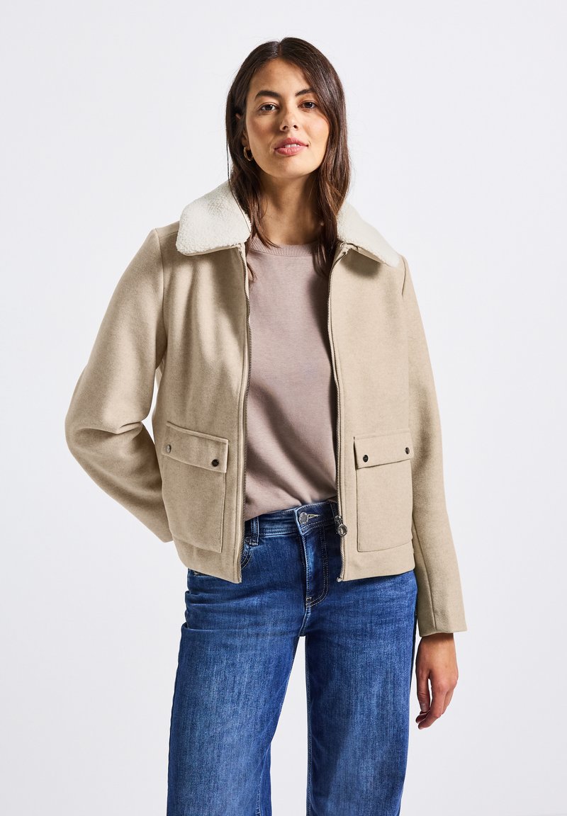 Žena v béžové zipové bundě s bílým fleecovým límcem, s mauve tričkem a modrými džínami, stojí proti jednoduchému bílému pozadí.