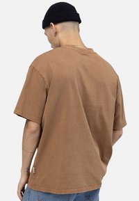 Man wearing brown oversized T-shirt and black beanie, facing away, showing back and left arm tattoo with light background.
