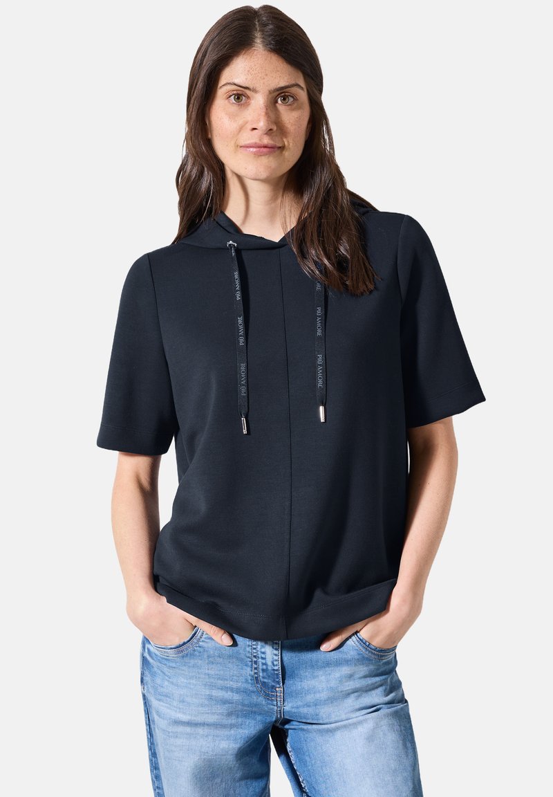 Sudadera negra de manga corta con cordones y puntas metálicas, combinada con denim azul claro. Tejido suave, corte holgado, detalles mínimos.