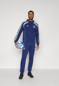 Mørkeblå treningsdrakt med lysegrønne striper, som har hettegenser og smale bukser. Inkluderer en sportsball i modellens hånd.