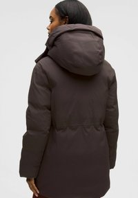 Doudoune marron foncé avec une grande capuche, taille cintrée et tissu lisse. Présente une coupe décontractée et sans matériel visible. Surface texturée.