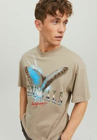 T-shirt beige a maniche corte con una grande grafica di una farfalla blu e il testo "ORIGINALS" in carattere metallico. Materiale in morbido cotone.