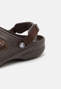 Sandale ajustable marron avec un dessus texturé, semelle en caoutchouc et grip à clous. Un pin's logo sur le côté et une sangle de talon rembourrée ajoutent des détails.