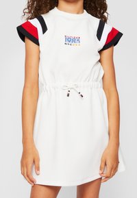 Weißes kurzes Kleid mit Kordelzug in der Taille, mit schwarzen, roten und marineblauen gestreiften Akzenten an den Ärmeln und gesticktem Logo auf der Brust.