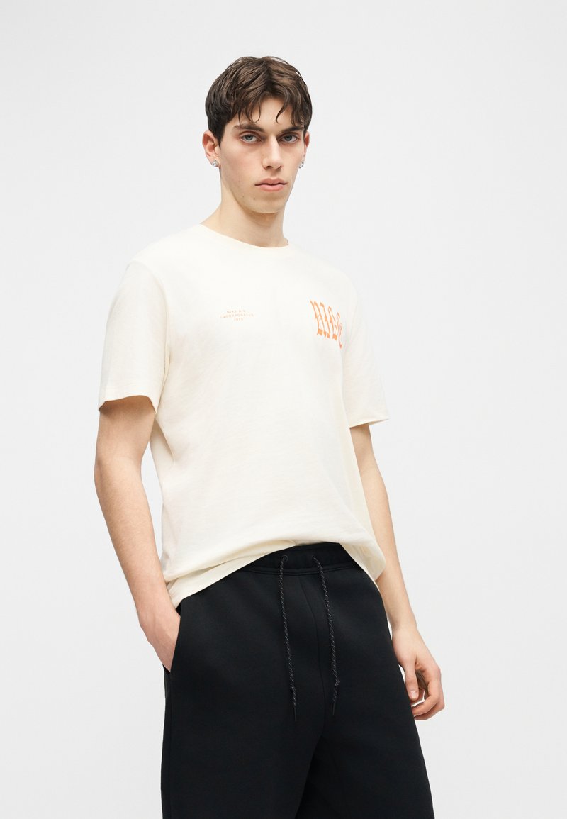 Jeune homme aux cheveux effet mouillé porte un T-shirt graphique blanc cassé et un pantalon noir à cordon, debout avec une main dans la poche sur fond blanc.