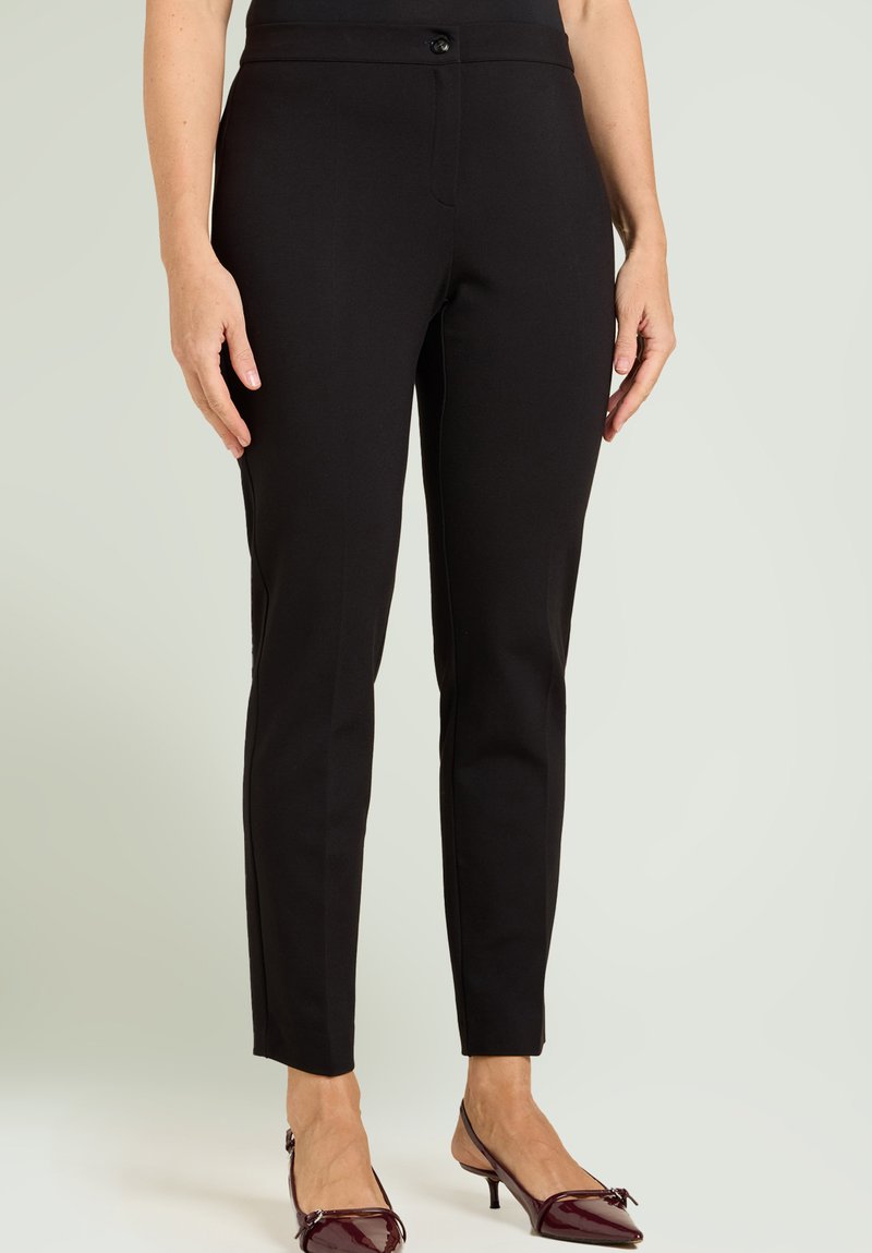 Donna che indossa pantaloni aderenti neri alla caviglia e décolleté slingback color bordeaux con punta affilata, in piedi davanti a uno sfondo semplice.