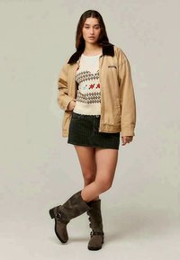 Casaco oversized bege com colarinho preto, suéter branco com padrões coloridos, saia mini preta com riscas e botas castanhas até ao joelho.