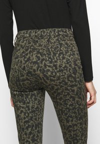 Jeans verde oliva con un patrón de leopardo negro; cuenta con dos bolsillos traseros y una cinturilla de altura media, fabricados con una tela suave y texturizada.