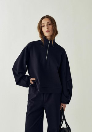 Kobieta ubrana w czarną, oversize'ową bluzę z przednim zamkiem oraz czarne spodnie, trzymająca czarną torbę na jednolitym tle.