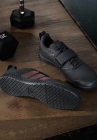 Para czarnych butów do podnoszenia ciężarów z paskiem na drewnianej podłodze obok dwóch hantli, z których jedna ma etykietę 25 funtów.