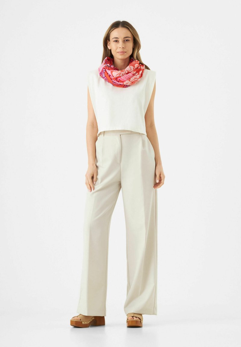 Een geknipte, mouwloze witte top gecombineerd met hooggerokte, wijde beige broek. Geaccentueerd met een kleurrijke sjaal met bloemenpatroon.